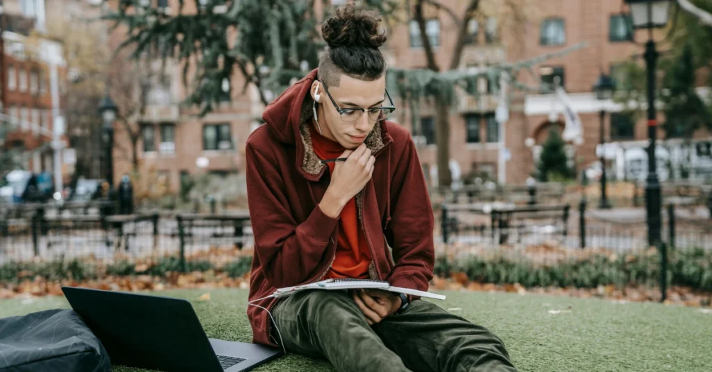 student with laptop and notebook sitting on grass in park